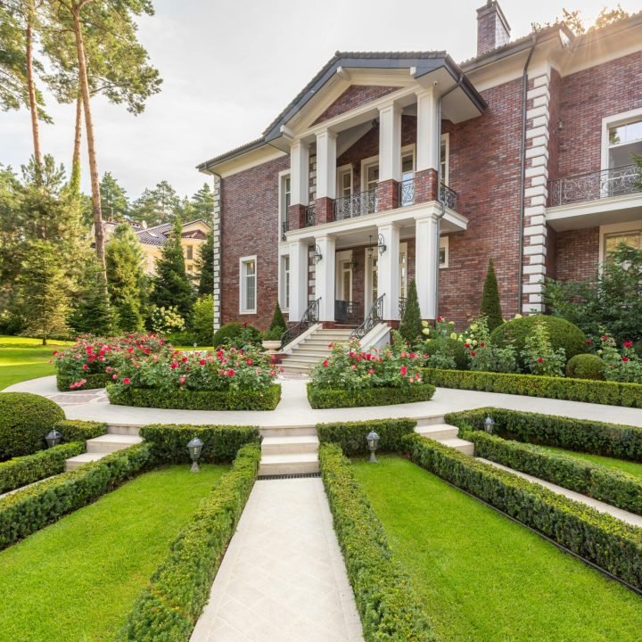 Luxurious brick mansion surrounded by lush gardens and trimmed hedges on a sunny day.